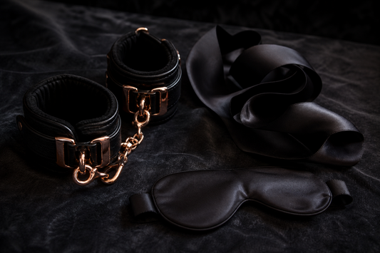 Black padded cuffs with rose-gold hardware, dark silk sash and satin blindfold on charcoal velvet — beginner bondage guide from Lace & Luna