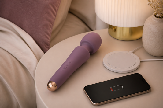 Rechargeable silicone wand vibrator on a cream bedside table beside a wireless charging pad and a smartphone showing a low battery indicator.